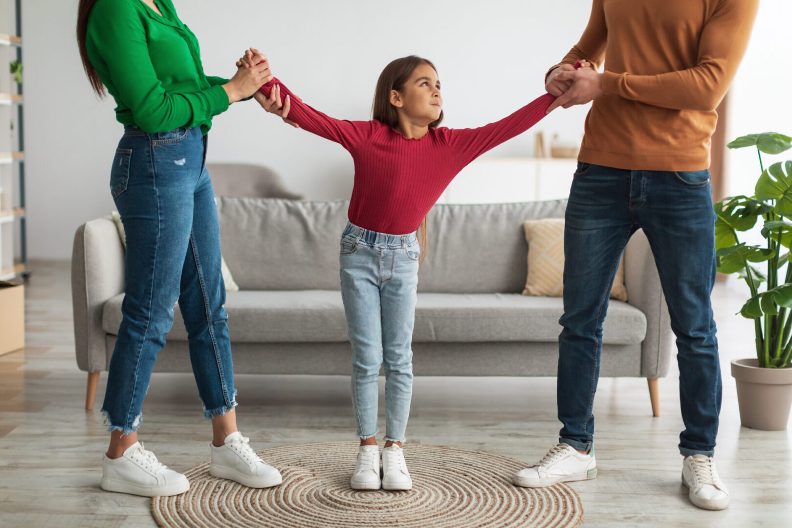 Divorce Concept. Angry parents fighting over their child, mad man and woman quarrelling, pulling daughter's hands in different directions standing in living room. Domestic Violence, Separation